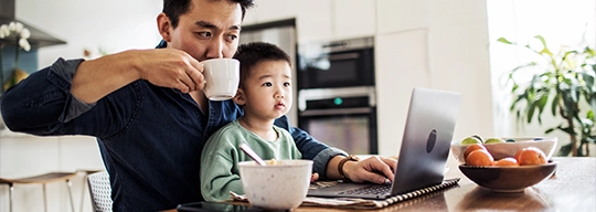 Un padre con su hijo en su regazo revisa su computadora portátil mientras toma una taza de café.